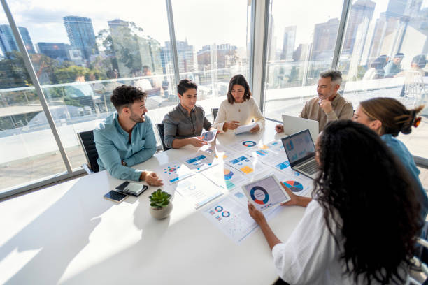 Multi racial group of people working with Paperwork on a board room table at a business presentation or seminar. The documents have financial or marketing figures, graphs and charts on them. There is a laptop and digital tablet  on the table. Multi ethnic group including Caucasian and African American